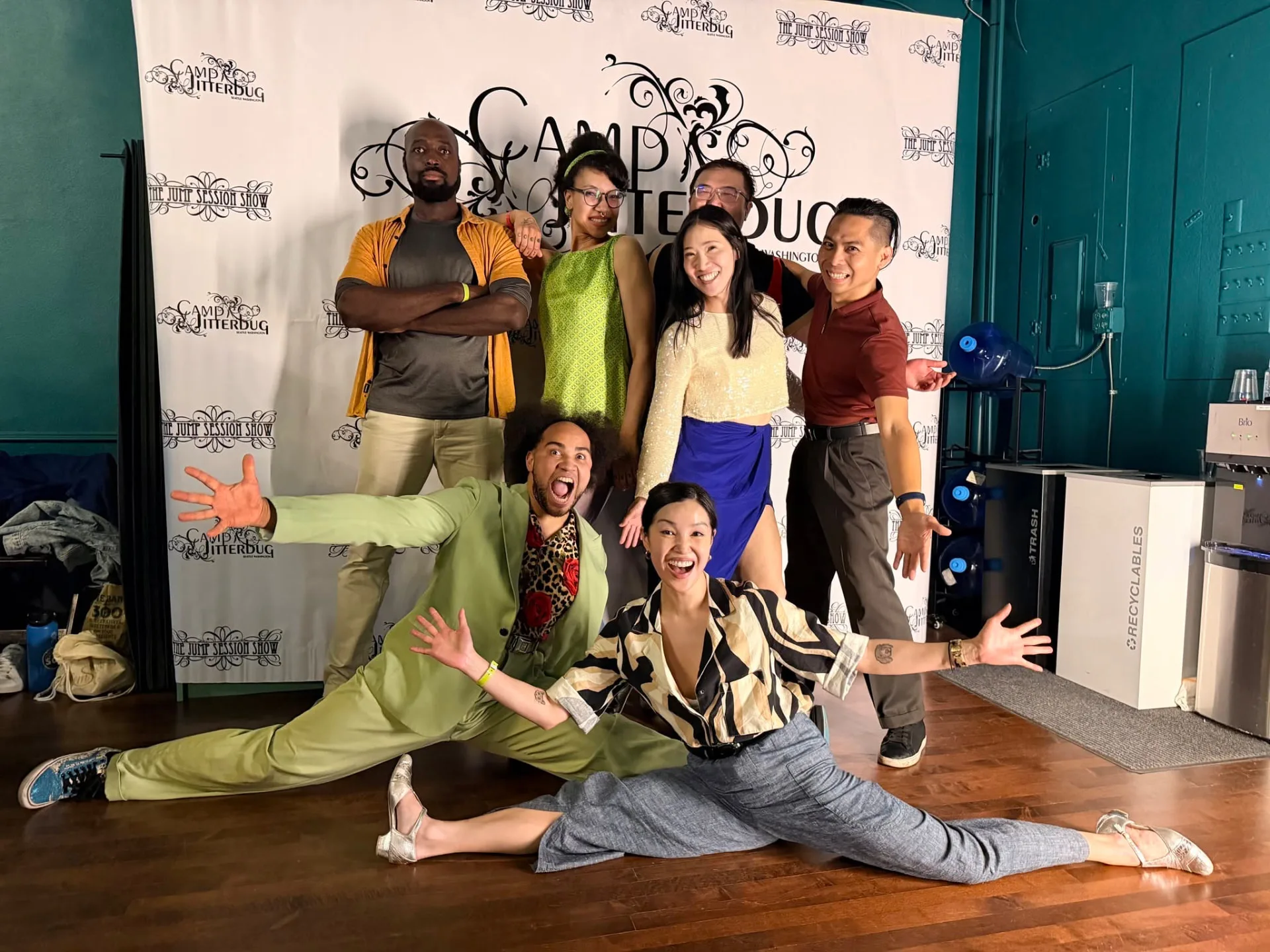 The Swing Arts Foundation performance group posed together in front of the Camp Jitterbug backdrop, two dancers in splits in front.